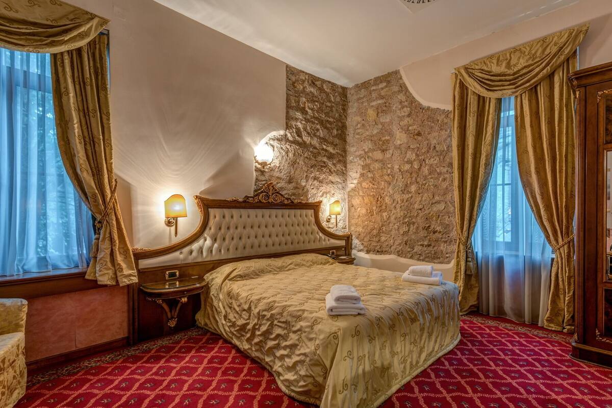 A luxurious hotel room with a large bed, ornate headboard, and a stone accent wall.