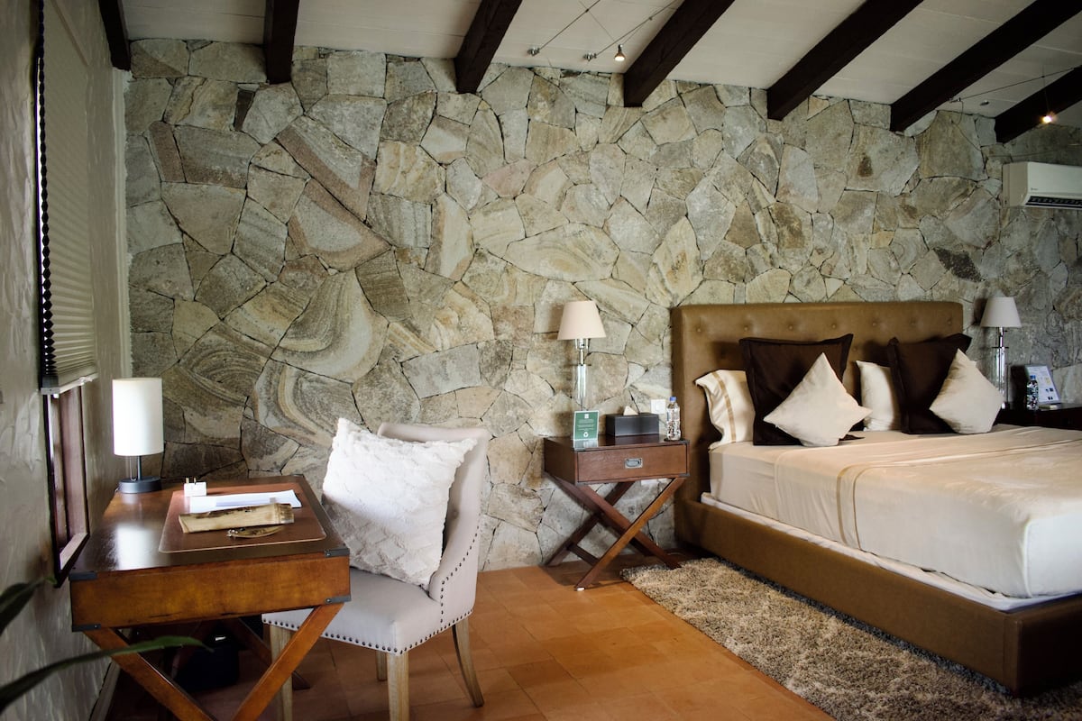 A bedroom with a stone wall, a wooden bed, a nightstand, a chair, and a desk with a lamp.