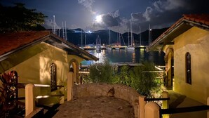 Una marina nocturna con barcos amarrados, un camino de piedra que conduce a un edificio y montañas al fondo.