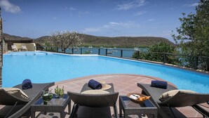 Zona junto a la piscina con sillones de mimbre, una mesa con bebidas y vistas a barcos en un lago.