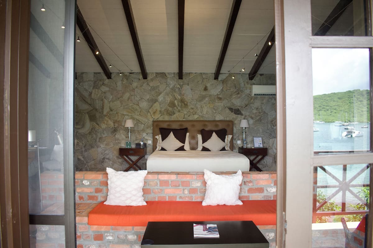 A bedroom with a stone wall, a bed with pillows, a nightstand, and a window with a view of boats and greenery.