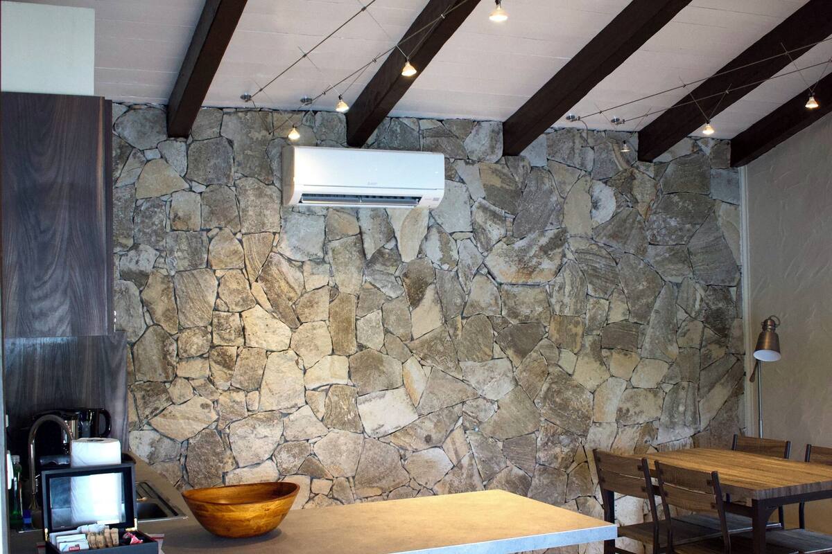 A modern kitchen with a stone wall, wooden beams, a large island with stools, and a dining area with chairs.
