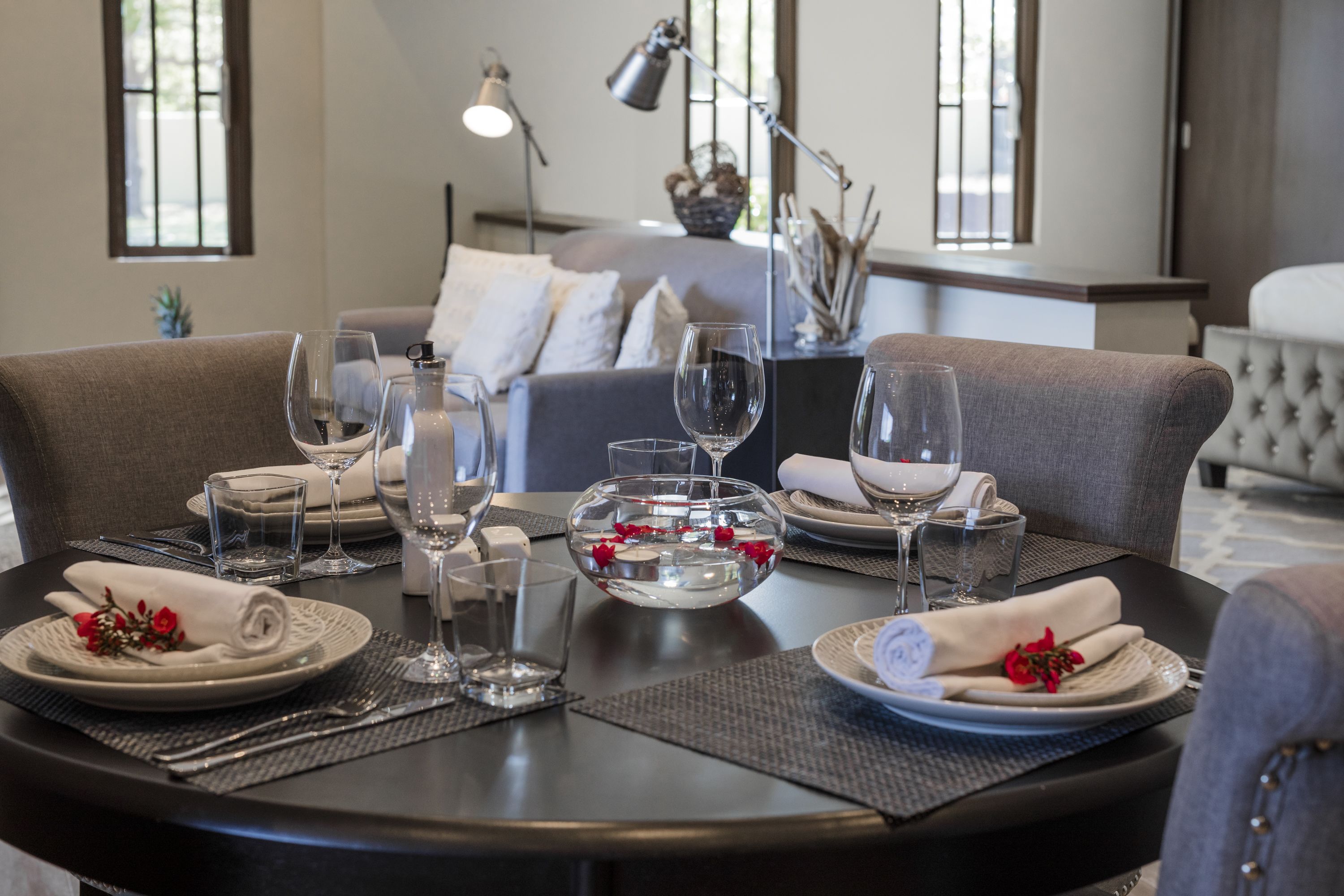 Una mesa de comedor dispuesta con platos, vasos y un cuenco decorativo, con un sofá y una lámpara al fondo.