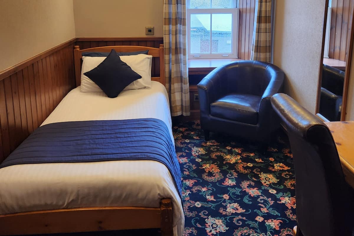 A hotel room with a bed, a chair, a desk, a window with curtains, and wall-mounted lights.
