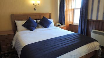 A neatly made bed with a white and navy blue bedspread, two navy blue pillows, a wooden headboard, a nightstand with a telephone, and a window with curtains.