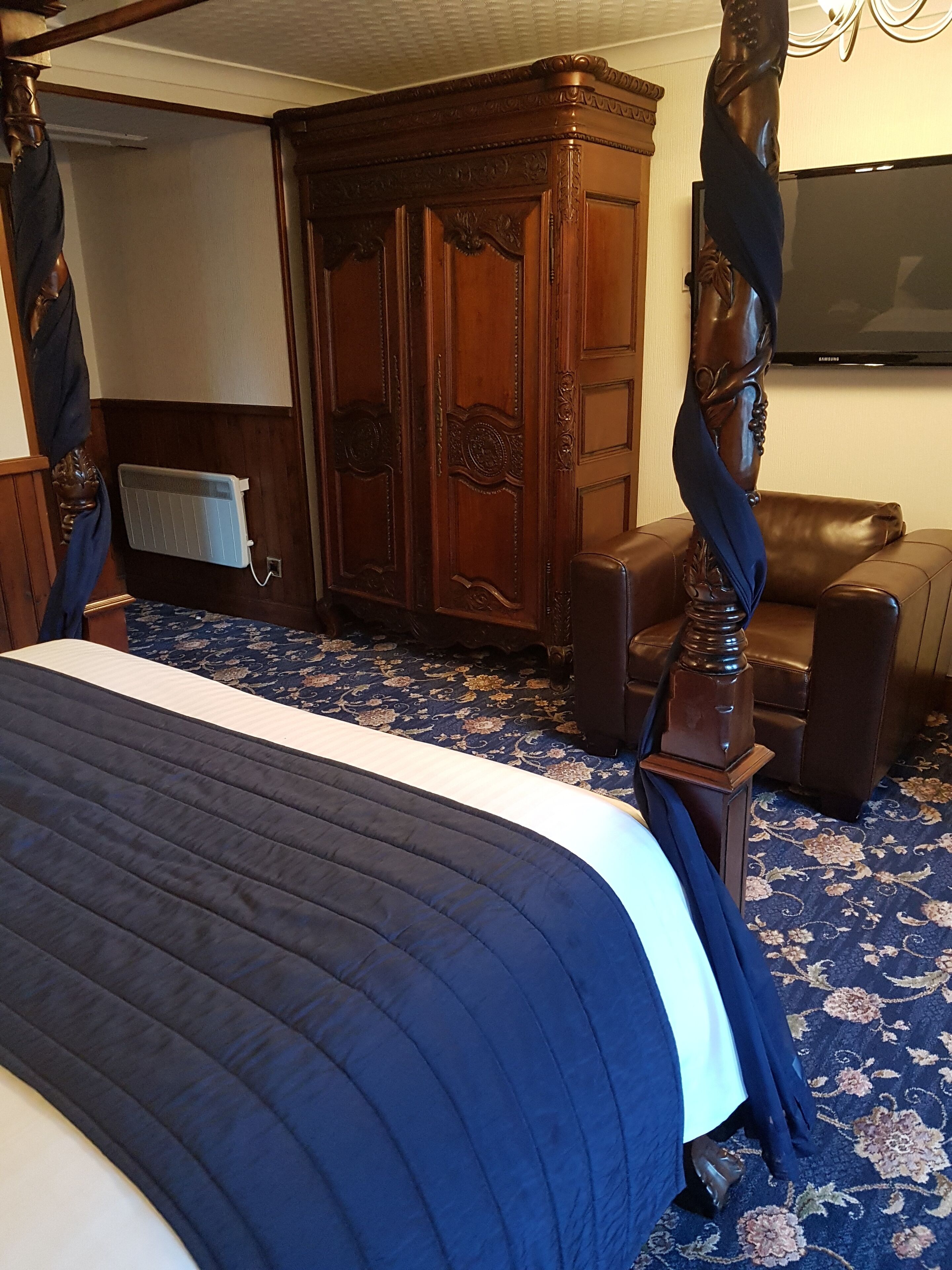 A bedroom with a large bed, a wooden wardrobe, a leather armchair, and a television on the wall.
