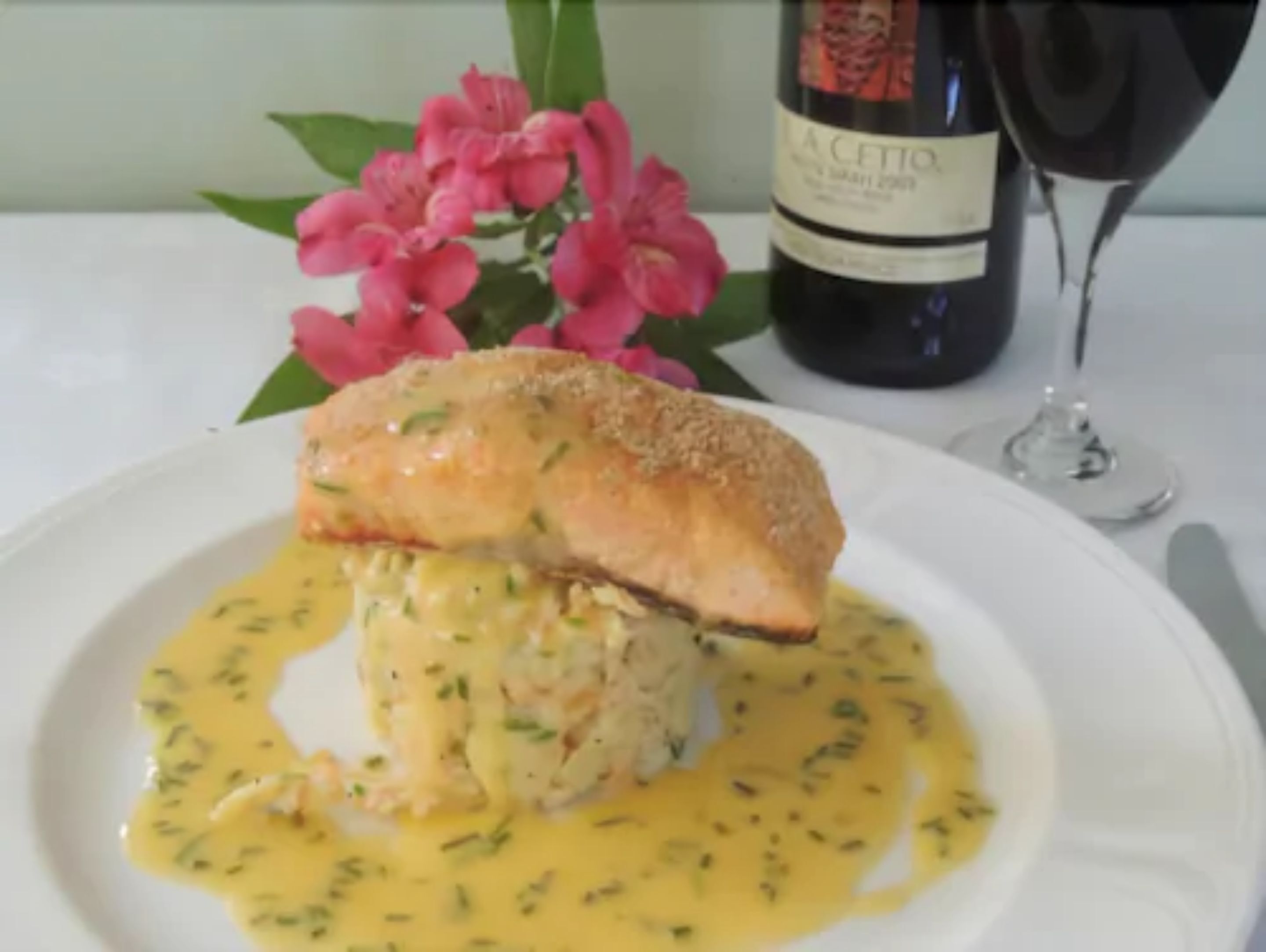 A plate with a breaded and fried item, possibly a fish fillet, served with a creamy sauce and garnished with herbs. A bottle of wine and a glass are visible in the background.