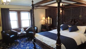 A four-poster bed with a dark wooden frame, a blue and white bedspread, and two blue pillows. There is a small table with a lamp and a chair in front of a window with curtains. - The Orkney Hotel (Kirkwall)