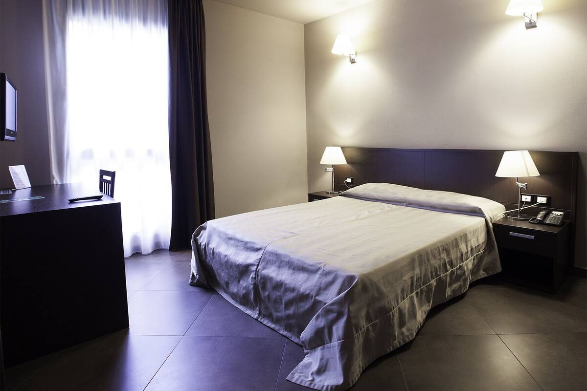 Ein Hotelzimmer mit einem großen Bett, zwei Nachttischen mit Lampen, einem Schreibtisch mit Stuhl und einem Fenster mit Vorhängen.