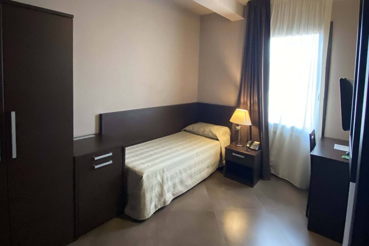 Ein Hotelzimmer mit Bett, Nachttisch mit Lampe, Schrank und einem Fenster mit Vorhängen.