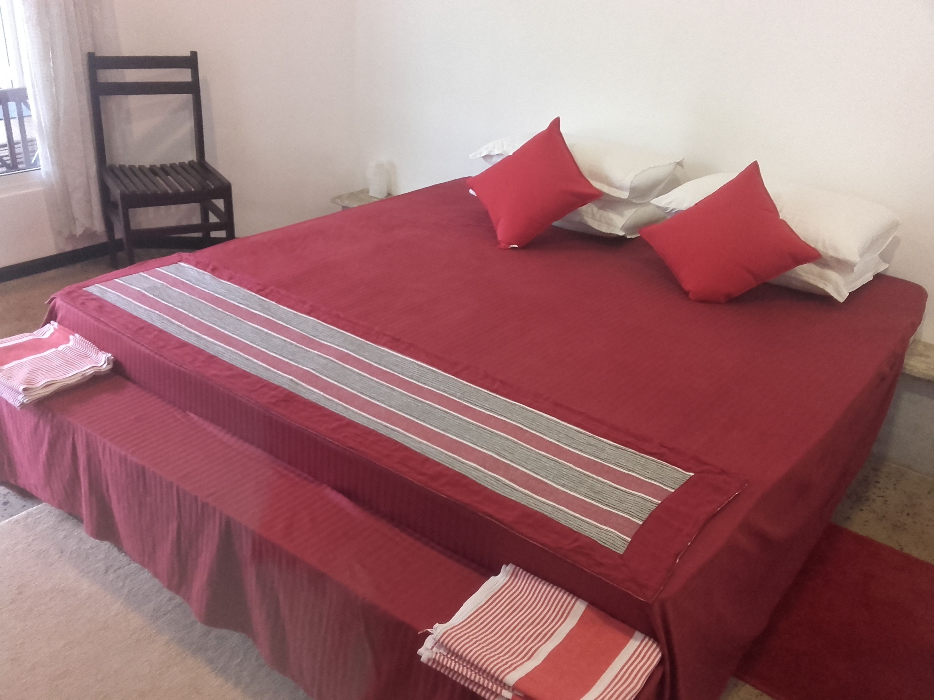 Una cama bien tendida con una colcha roja, dos almohadas rojas y una almohada a rayas sobre una funda de almohada blanca.
