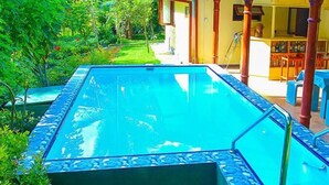 Piscina retangular com borda azulejada, rodeada por um jardim e um edifício com área de bar.