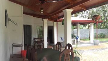 Un área de comedor al aire libre techada, con un ventilador de techo, sillas de madera y una mesa con mantel verde.