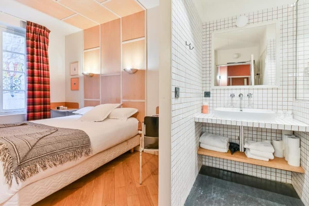 A bedroom with a bed, bedside tables, a window with curtains, and a bathroom with a sink, mirror, and towel rack.