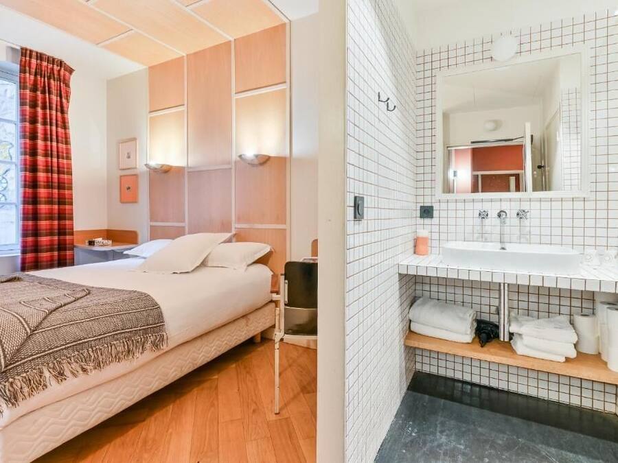 Une chambre à coucher comprenant un lit, des tables de chevet, une fenêtre avec des rideaux, ainsi qu’une salle de bain avec un lavabo, un miroir et un porte-serviettes.