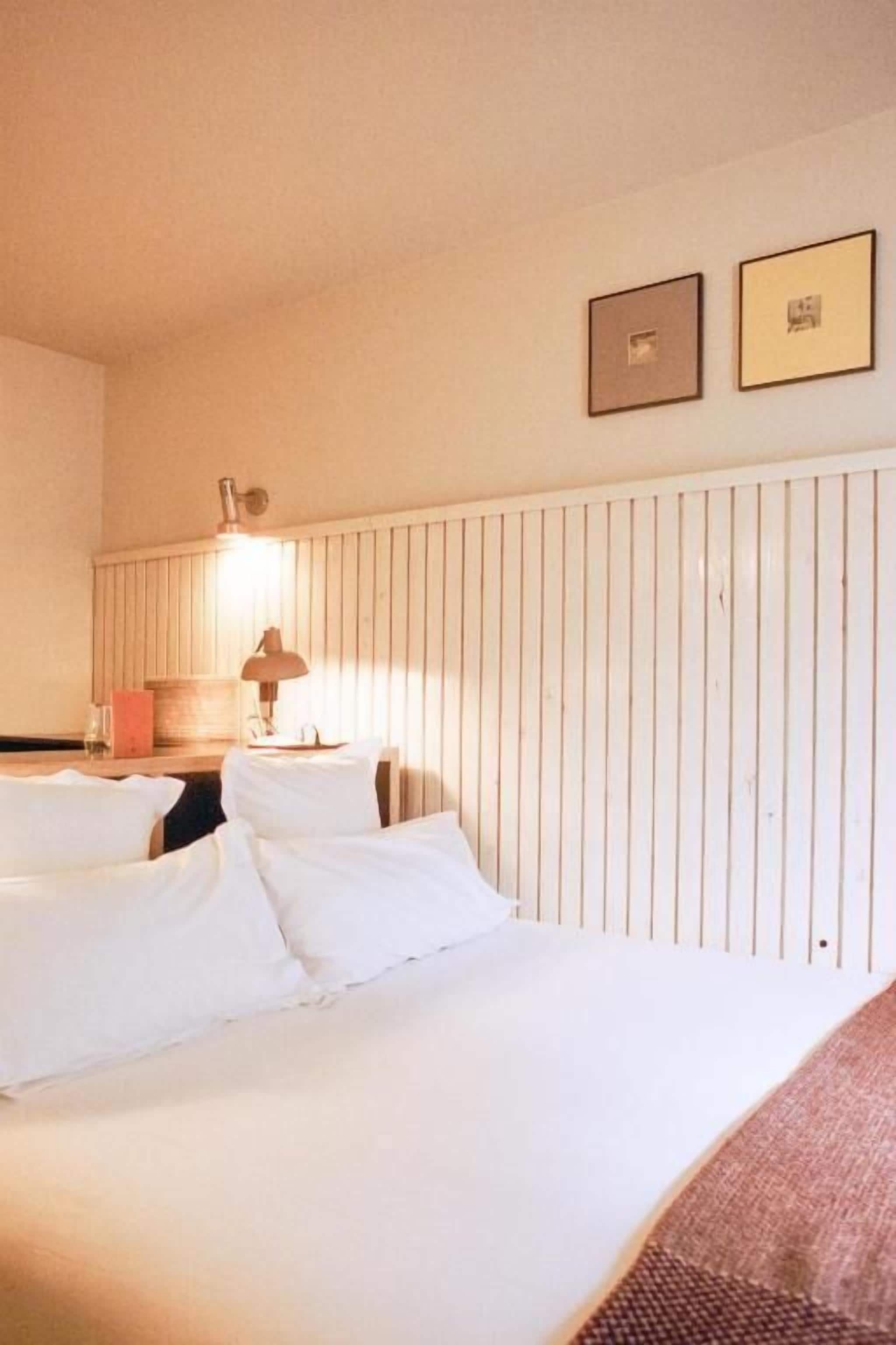 A hotel room with two beds, a wooden headboard, a lamp, and two framed pictures on the wall.