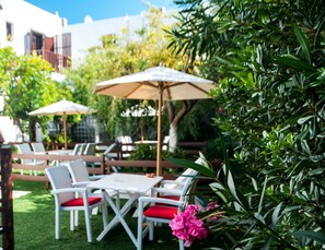 An outdoor seating area with white tables and chairs, some with red cushions, and beige umbrellas. - Maestralia (Skiros)