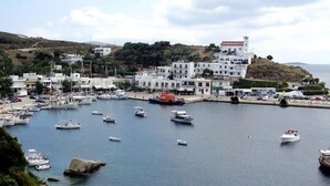 Uma vila costeira com um porto cheio de barcos, edifícios brancos com detalhes em azul, e um cenário montanhoso ao fundo.