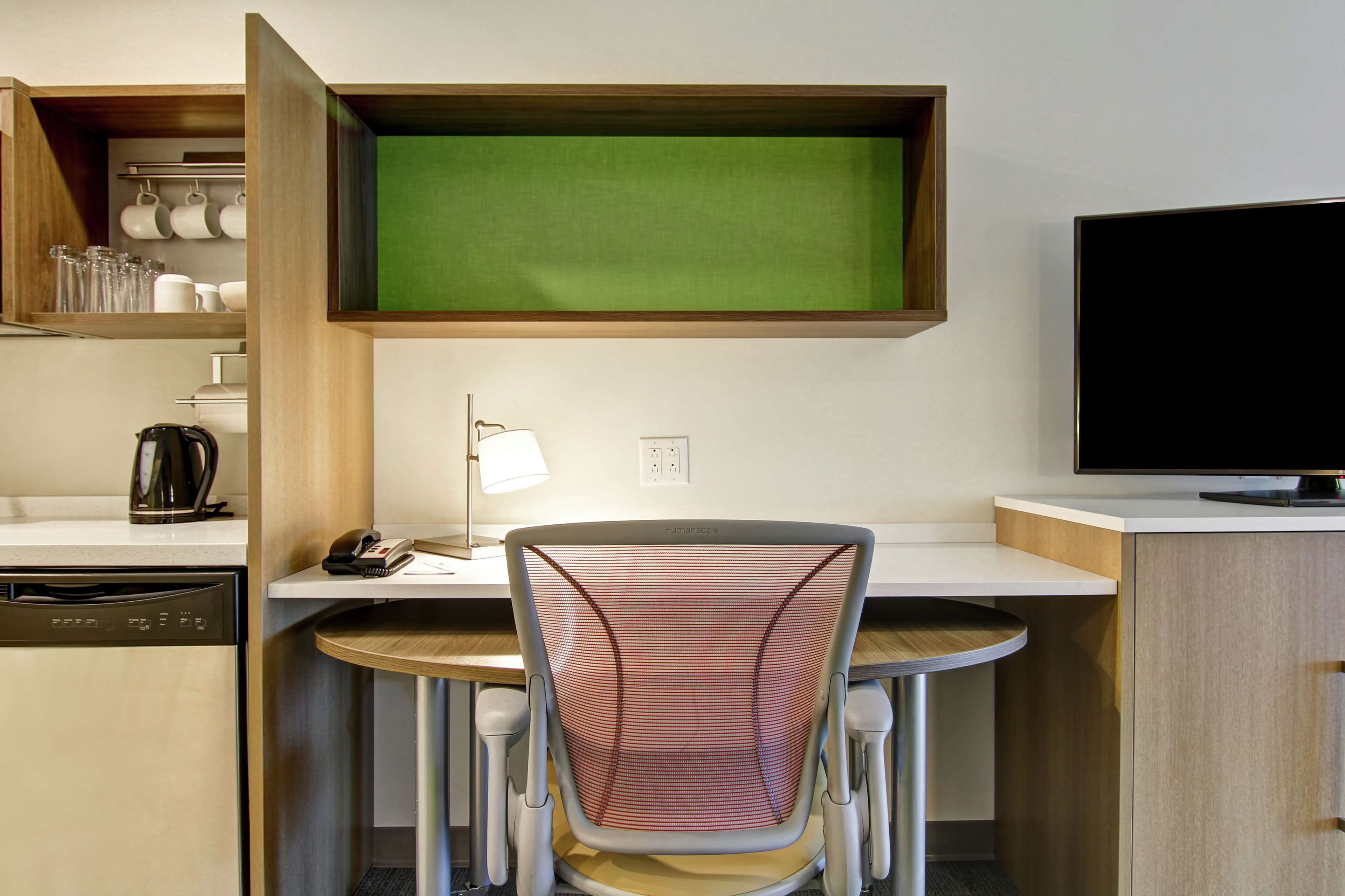 Habitación de hotel compacta con kitchenette, un comedor con una silla, televisor y pared verde tipo pantalla.