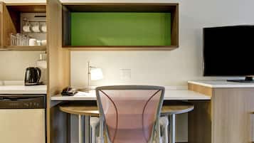 Habitación de hotel compacta con kitchenette, un comedor con una silla, televisor y pared verde tipo pantalla.