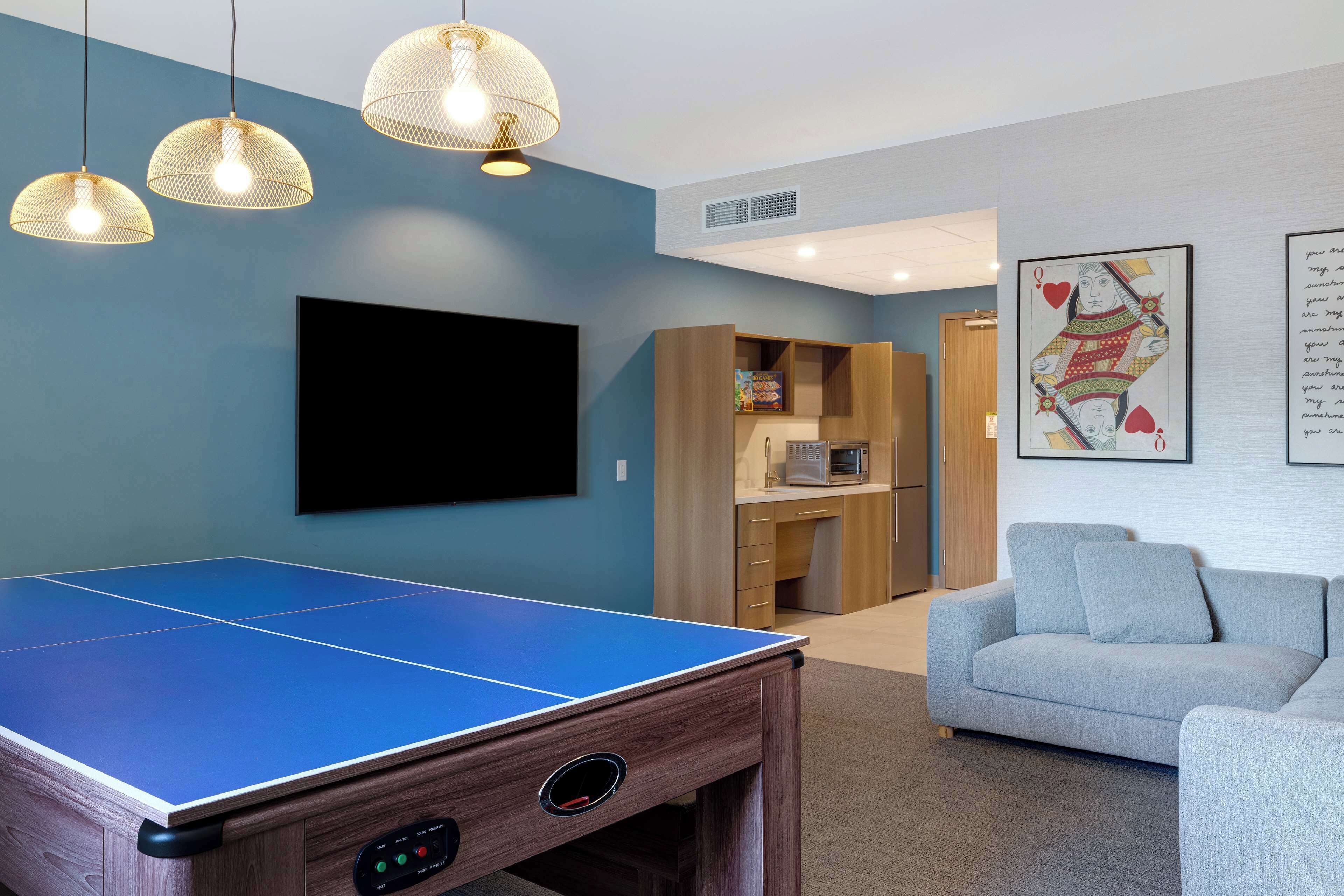A room with a ping pong table, a flat-screen TV, a sofa, and a kitchen area.