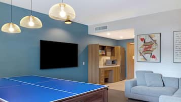 A room with a ping pong table, a flat-screen TV, a sofa, and a kitchen area.