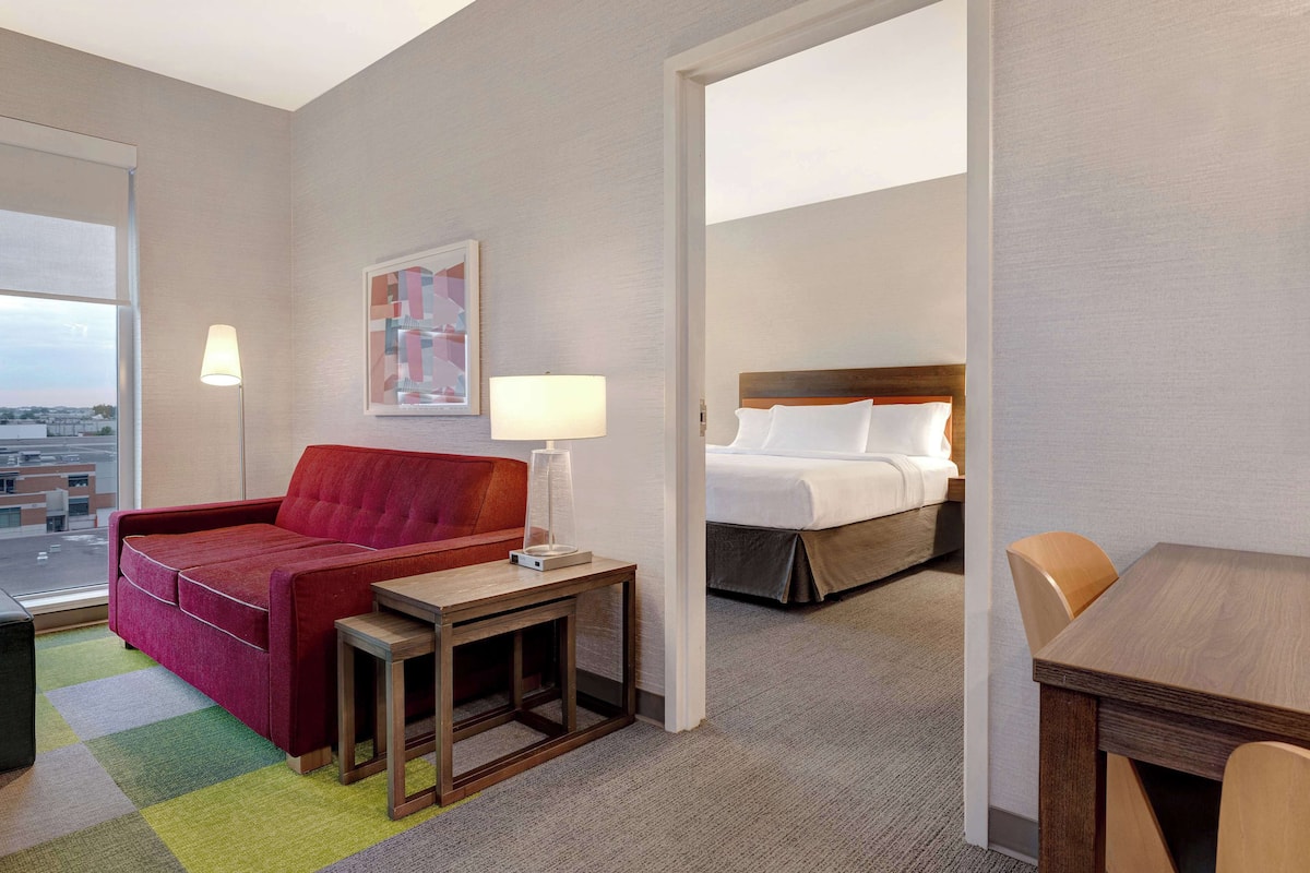Hotelkamer met rode bank, een klein tafeltje, lamp, bed met witte lakens en uitzicht door een groot raam.