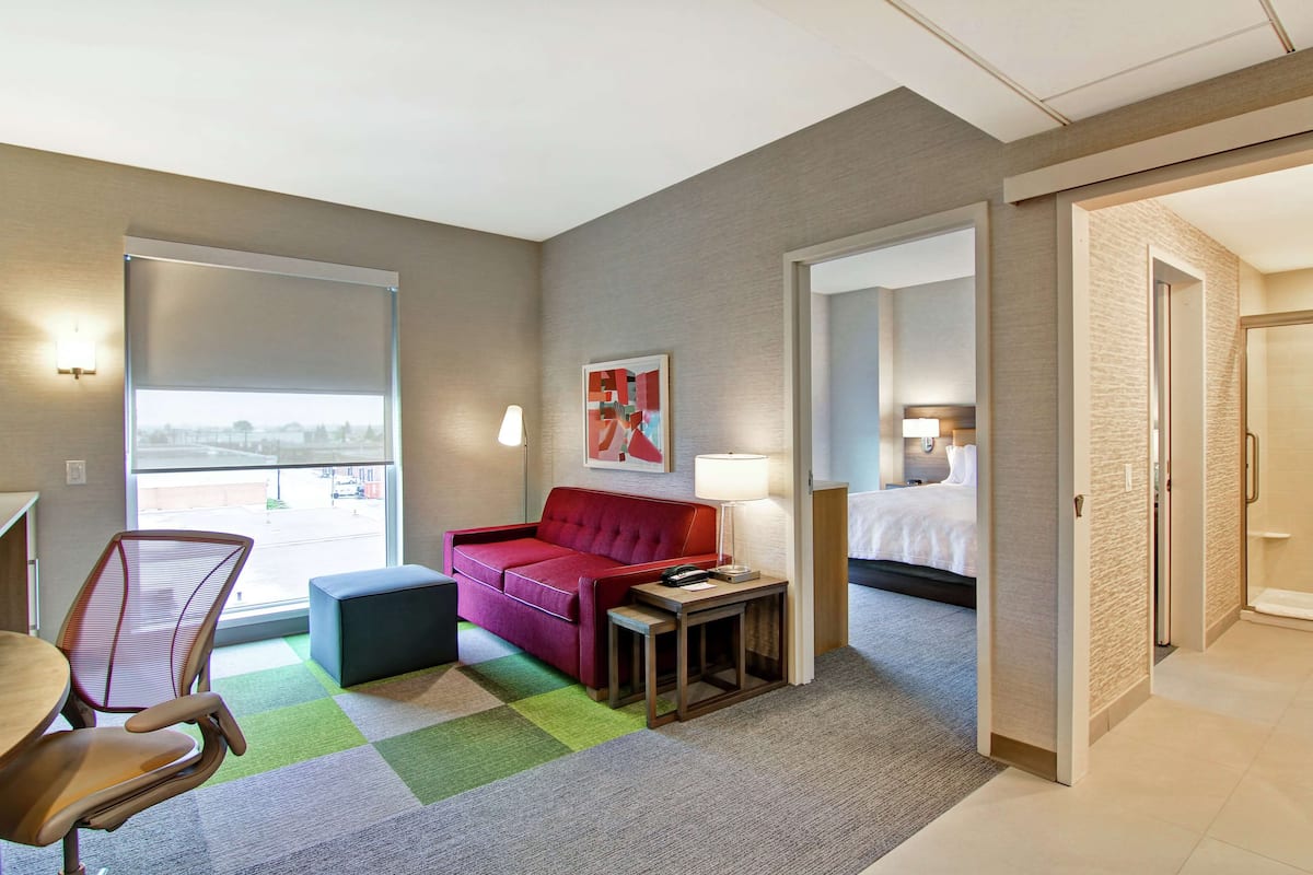 Hotelkamer met een rode bank, een tafeltje met een lamp, een stoel, een bed en uitzicht op de straat door een groot raam.