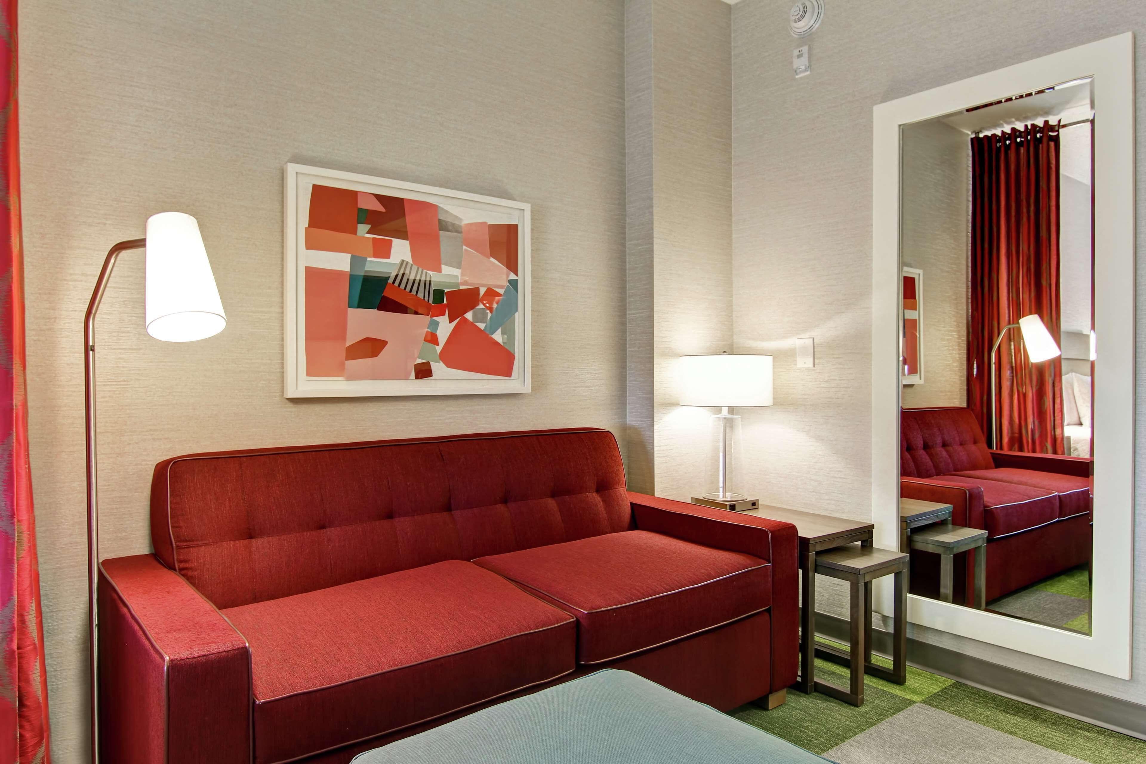 A modern hotel room with a red sofa, a small table, a lamp, and a framed abstract painting on the wall.