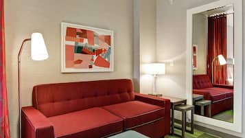 A modern hotel room with a red sofa, a small table, a lamp, and a framed abstract painting on the wall.