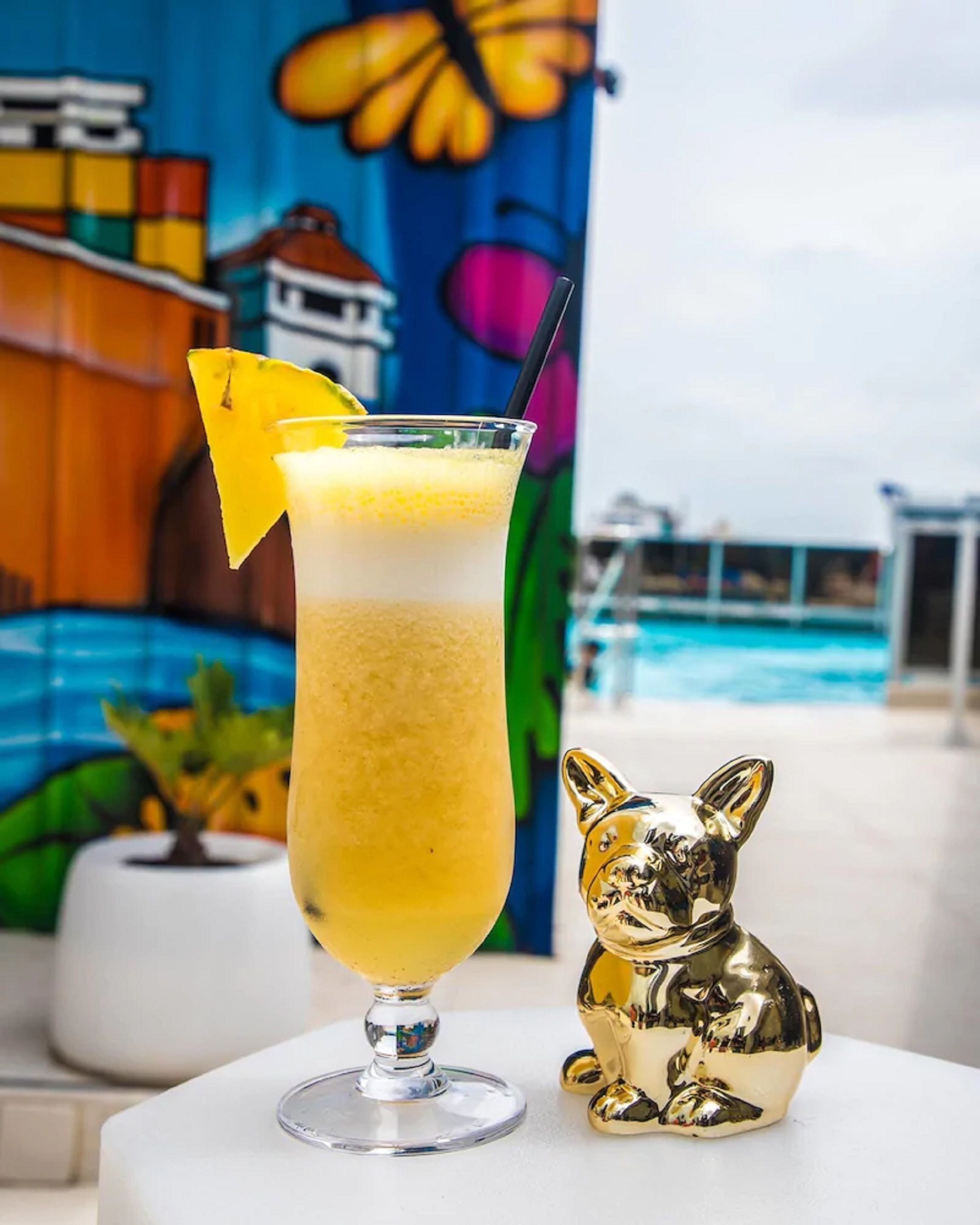 A tall glass cocktail with a layered drink, a gold bunny figurine, and a colorful mural in the background.