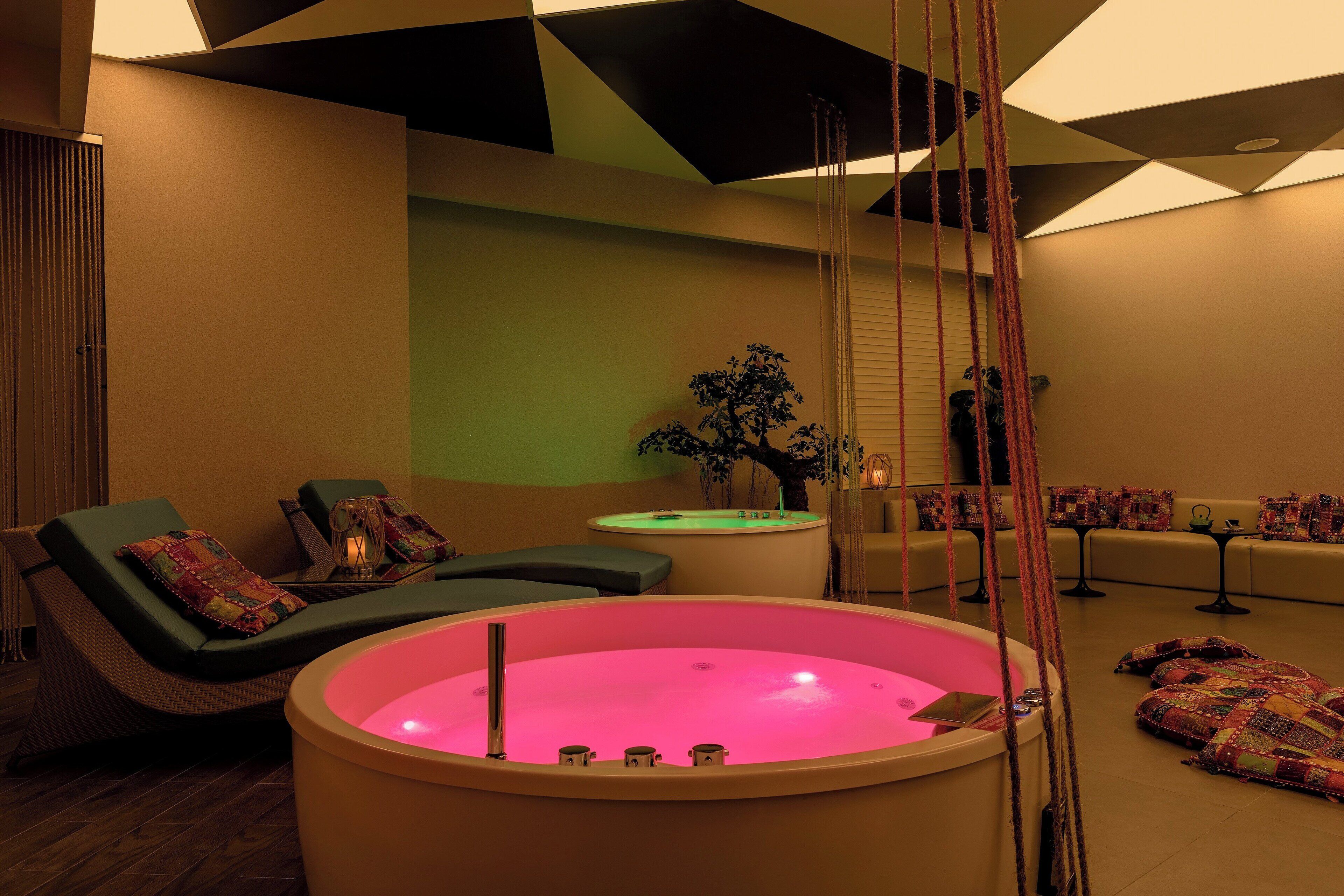 Une salle de spa dotée d’un grand jacuzzi rose, de fauteuils verts et d’une petite table sur laquelle est posée une plante.