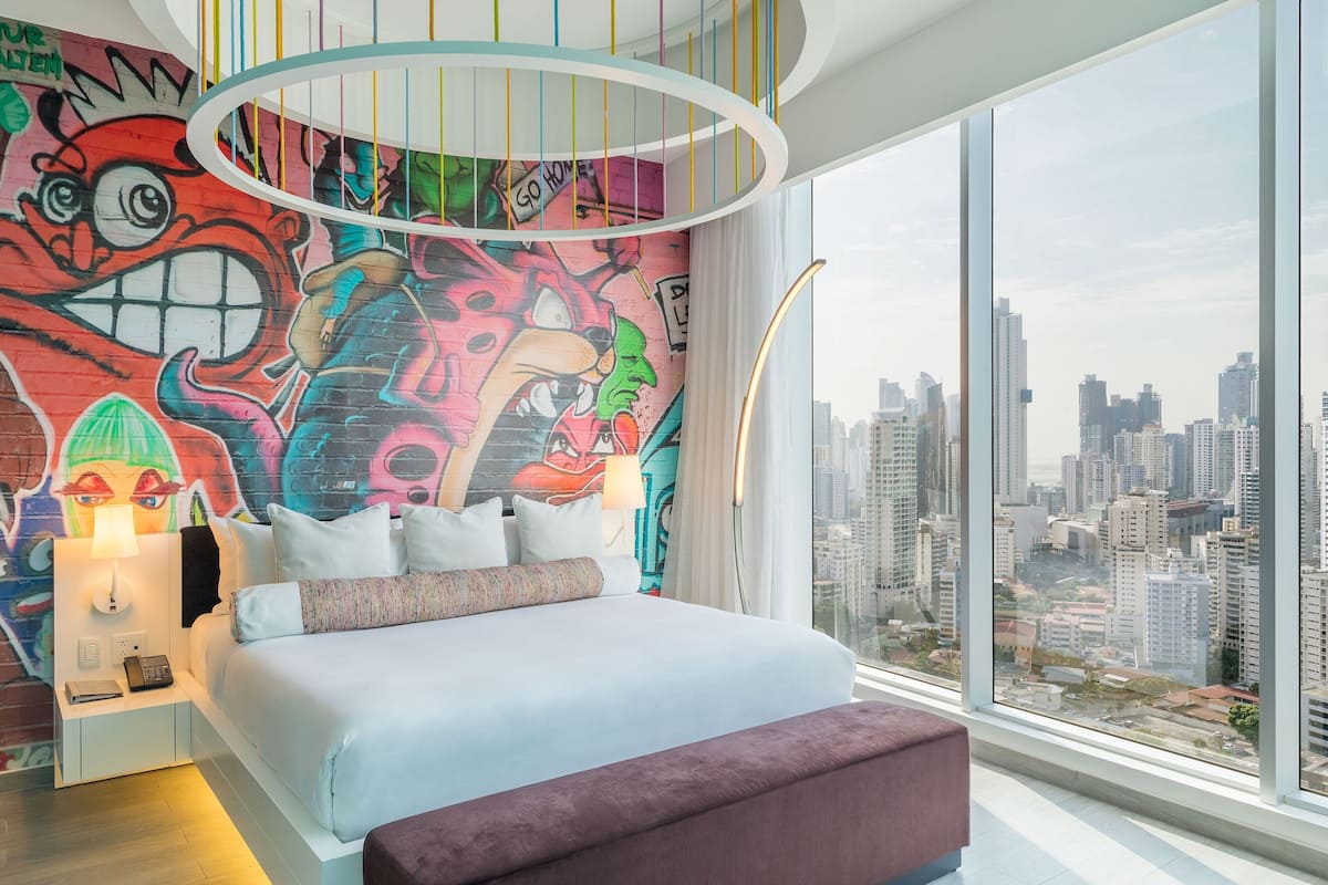 Habitació d'hotel moderna amb un llit gran, un mural colorit i vistes a la ciutat a través de grans finestres.