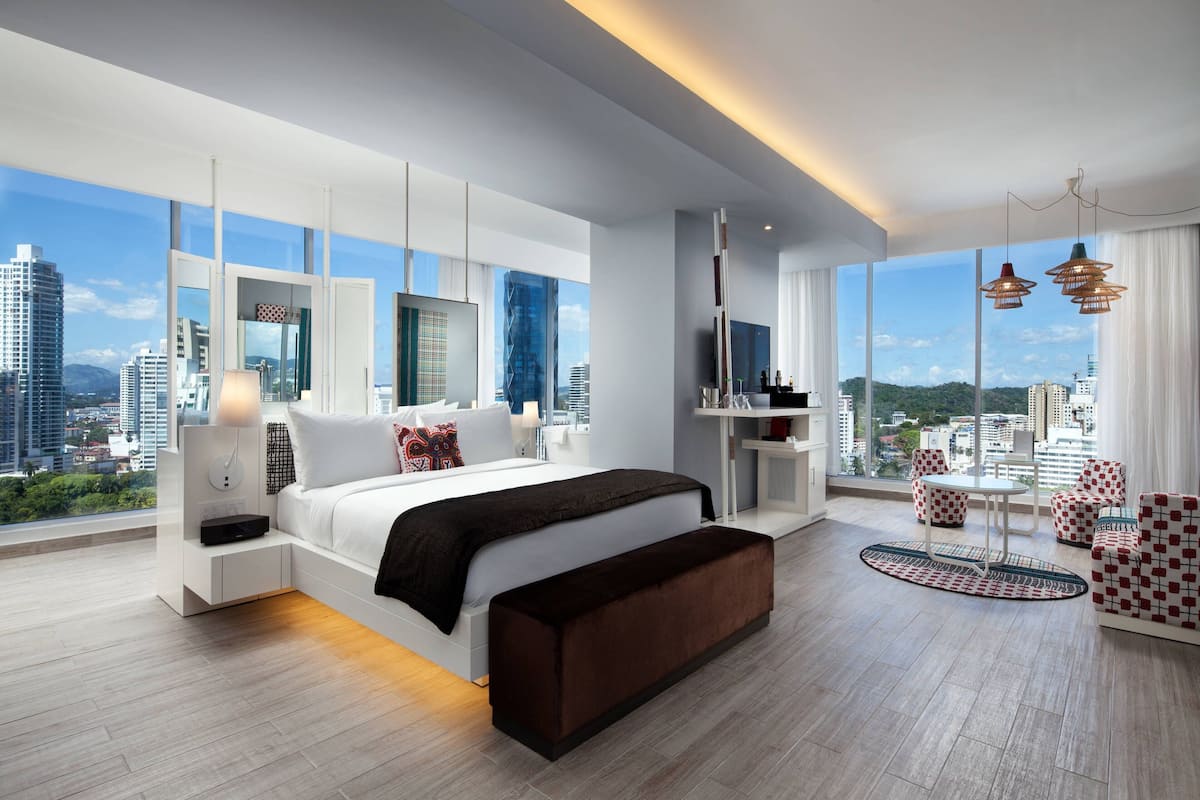 Habitació d'hotel moderna amb un llit gran, una zona de seients amb cadires, una taula petita i vista a la ciutat.