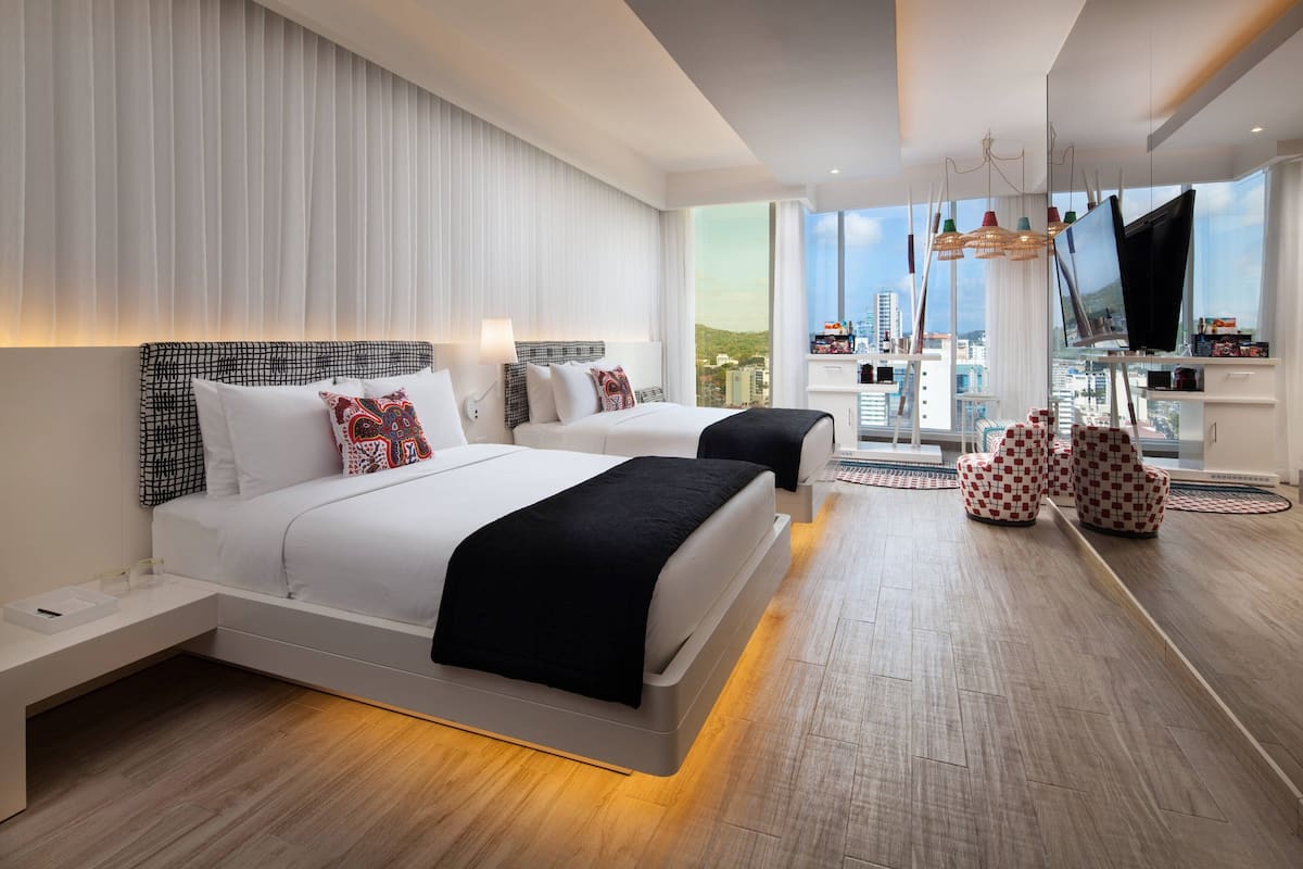 Una habitació d'hotel moderna amb dos llits, un gran finestral amb vista a la ciutat i una televisió sobre un suport.