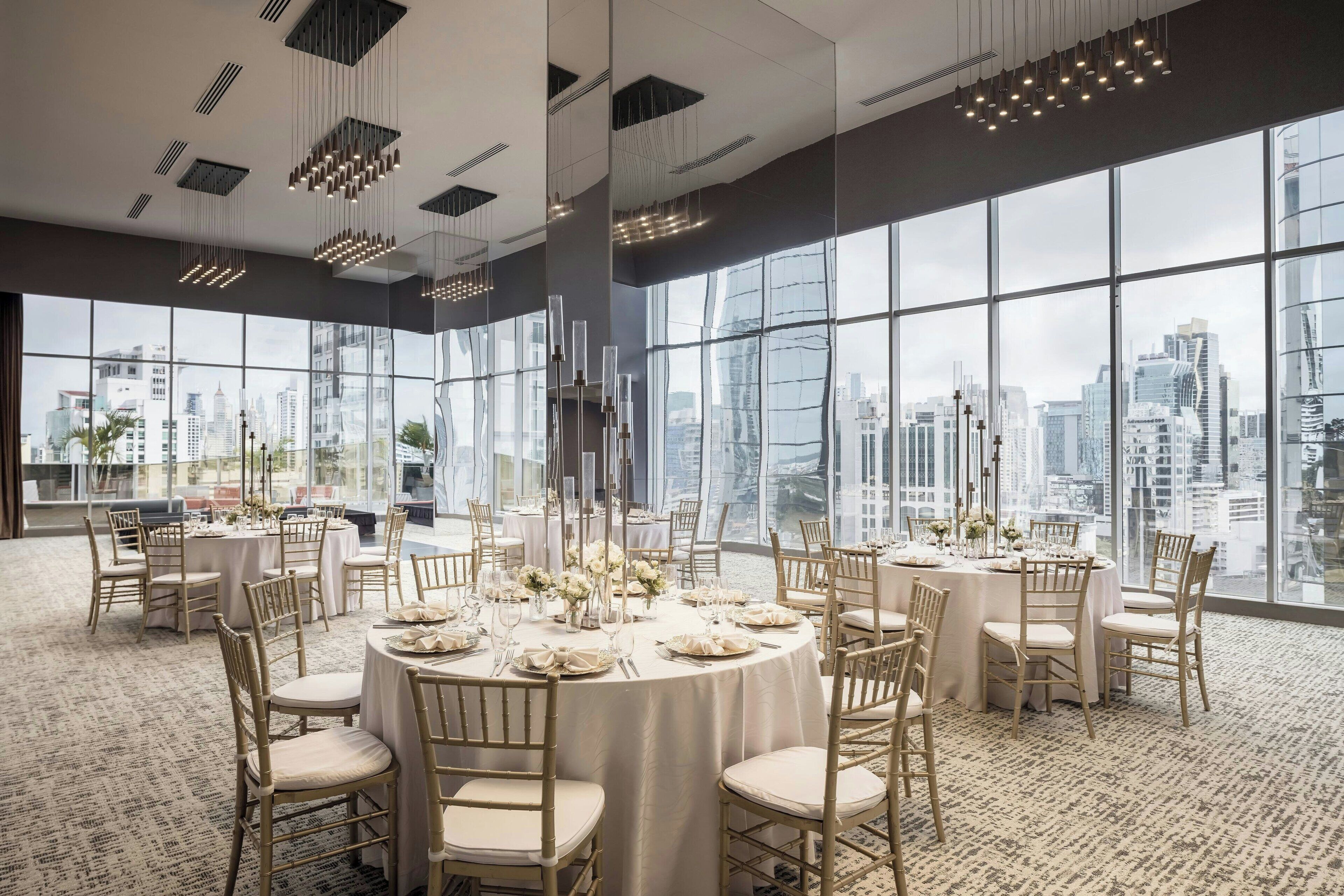 A high-rise hotel room with large windows offering a city view, round tables set for dining, and modern chandeliers.
