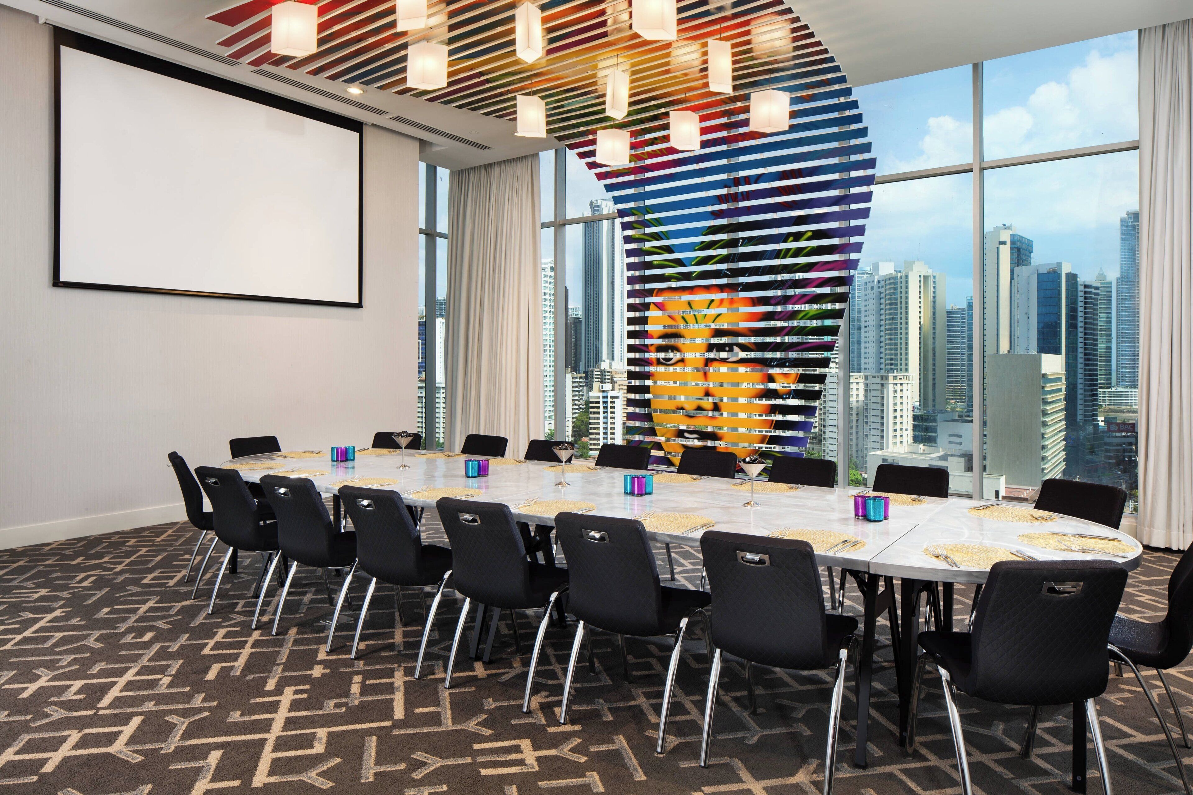 A conference room with a large table, black chairs, a projector screen, and a city view through large windows.