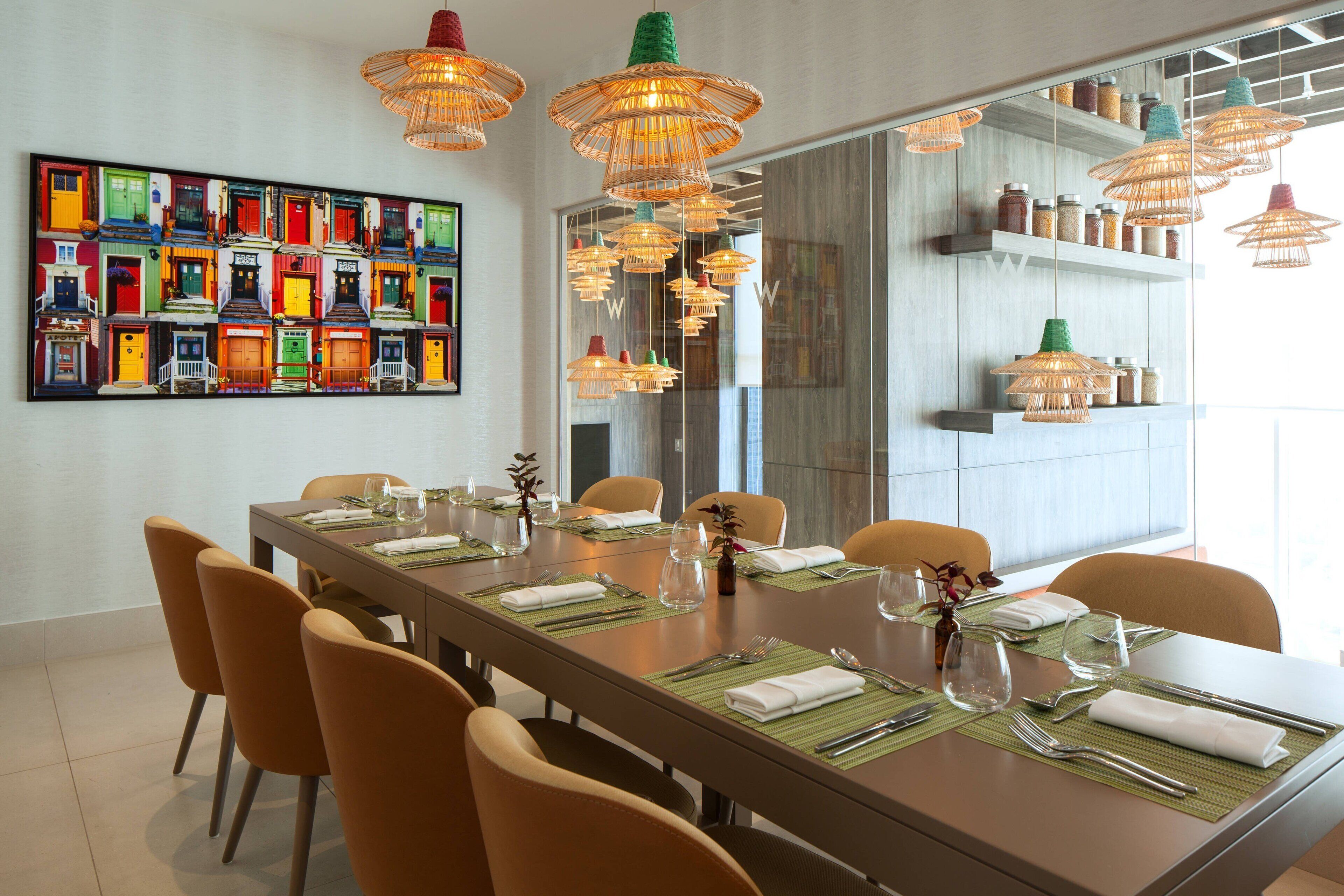 Un coin repas avec une longue table dressée pour six personnes, entourée de chaises orange, agrémenté d’une œuvre d’art colorée au mur et de luminaires suspendus en osier au-dessus.