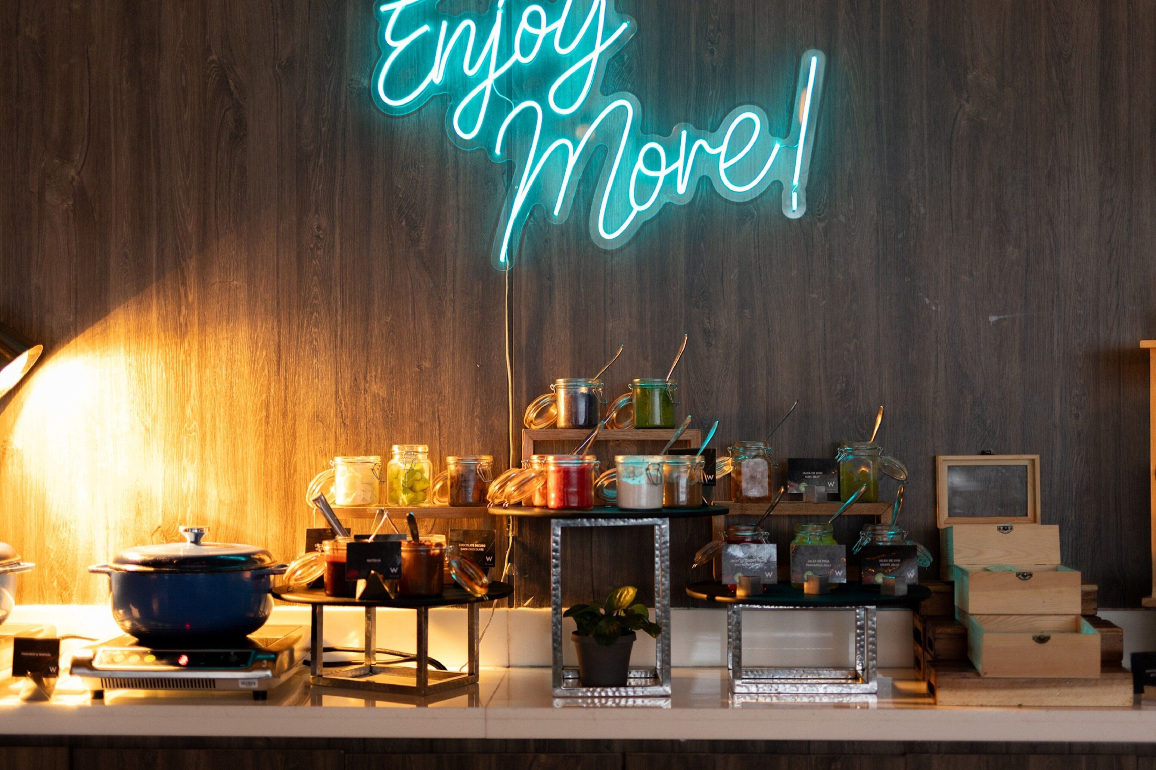 Una encimera de cocina con diversos utensilios de cocina, frascos y un letrero de neón que dice "Enjoy More!".