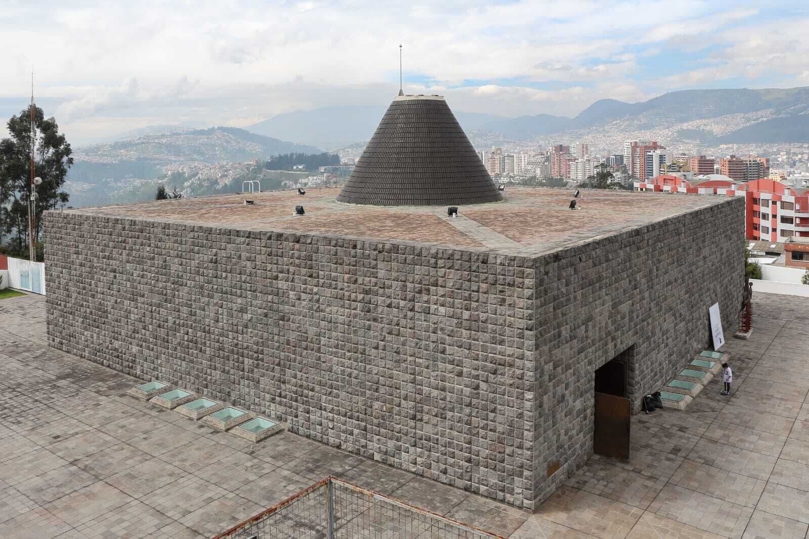 A large, square brick building with a conical roof, situated on a flat rooftop with a cityscape in the background.