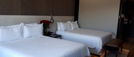 A hotel room with two beds, a nightstand, a vase with red flowers, and a bench.
