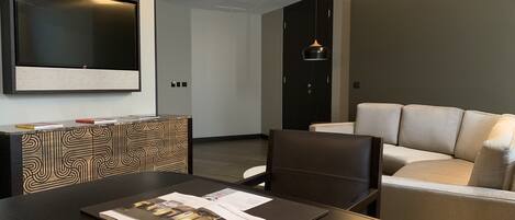 Un moderno cuarto de hotel con una mesa de comedor de madera oscura, una silla de cuero negro, un sofá color beige y un televisor de pantalla plana montado en la pared.