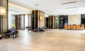 Un moderno vestíbulo de hotel con recepción, zona de estar y puertas de vidrio que dan a otras habitaciones.