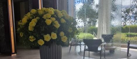 A hotel lobby with a large potted plant, a reception desk, and a glass wall with a view of the outdoors.