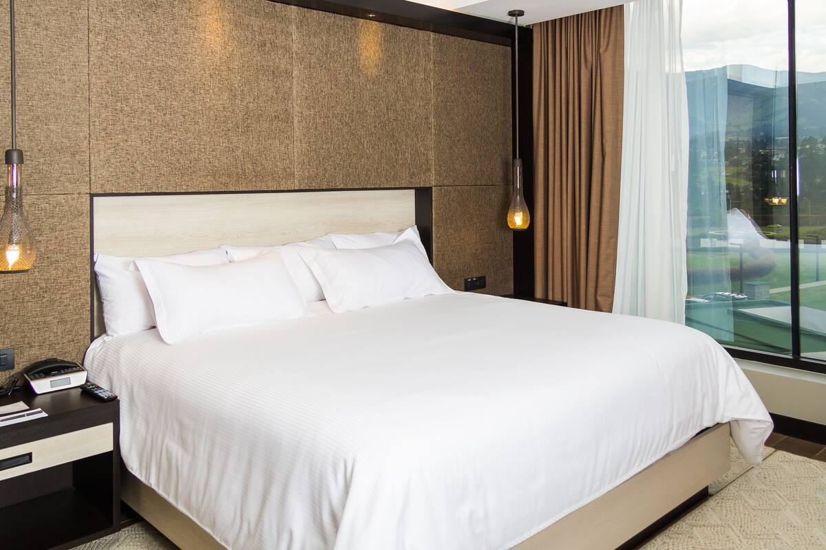 Habitación de hotel con una cama grande, una mesita de noche con teléfono y control remoto, una lámpara de pared y vistas a un paisaje urbano a través de ventanas amplias.