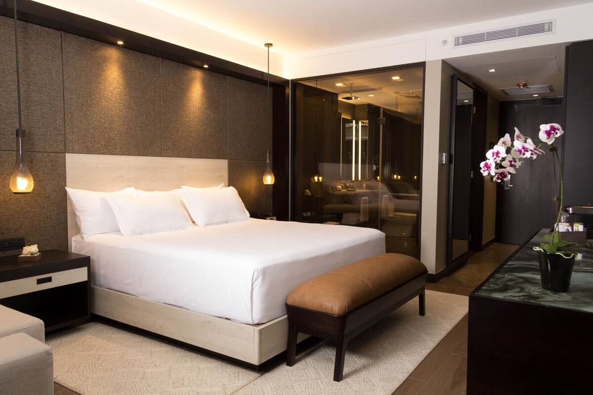 Una habitación de hotel moderna con una cama grande, mesitas de noche, un banco, un escritorio y un espejo.