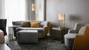 Ein modernes Hotelzimmer mit einem grauen Sofa, einem passenden Hocker, einem kleinen Beistelltisch und einer Lampe.
