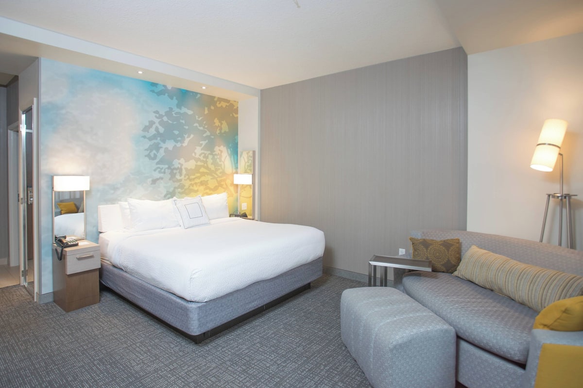 A modern hotel room with a large bed, a sofa, a small table, and a painting on the wall.