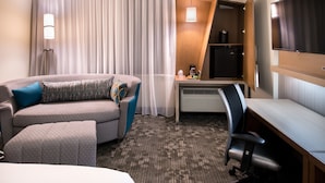 Ein Hotelzimmer mit Bett, Sofa, Schreibtisch, Stuhl, Fernseher und einem Fenster mit Vorhängen.