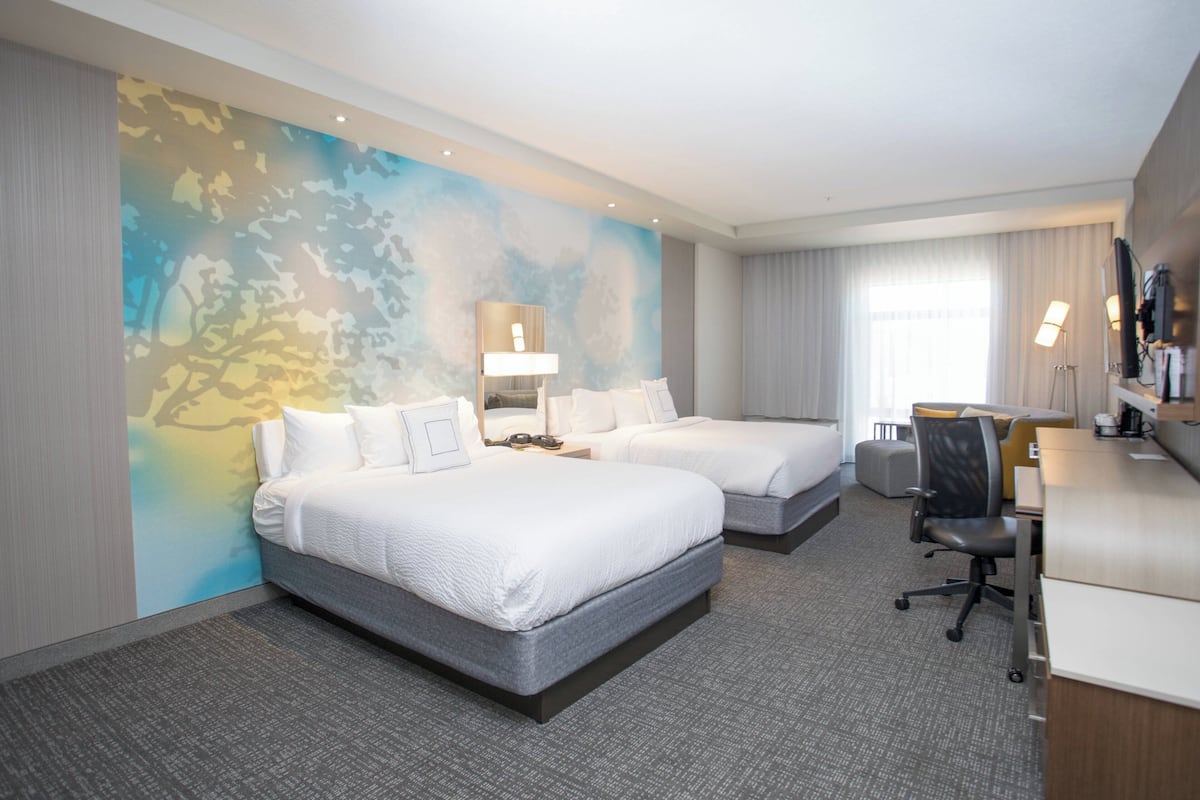 Ein Hotelzimmer mit zwei Betten, einem Schreibtisch, einem Stuhl und einem großen Fenster.